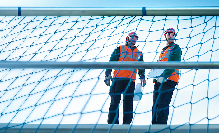 Zwei Personen stehen auf einem Gerüst, das mit einem Schutznetz gesichert ist.